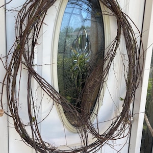 May include: A large, circular wreath constructed from thin, brown, intertwined natural twigs and branches. The wreath features an open, airy design with visible gaps between the woody stems, showcasing a raw, organic texture and dark brown color.