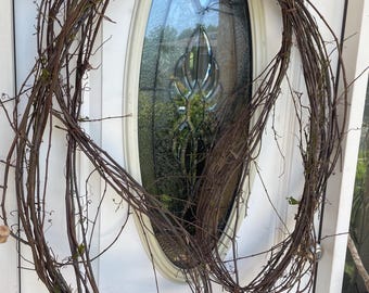 May include: A large, circular wreath constructed from thin, brown, intertwined natural twigs and branches. The wreath features an open, airy design with visible gaps between the woody stems, showcasing a raw, organic texture and dark brown color.