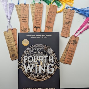 May include: A book titled "Fourth Wing" by Rebecca Yarros with a black cover and gold lettering. The book cover features a dragon with a circular design around it. The book is surrounded by wooden bookmarks with various quotes and designs.