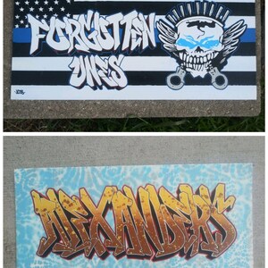 May include: Two hand-painted canvases with graffiti-style lettering. The top canvas features a blue and black American flag with a white stripe and the words "Forgotten Ones" in black and red. A skull with wings and handcuffs is in the upper right corner. The bottom canvas features a blue background with a white pattern and the word "ALEXANDERS" in gold and red.