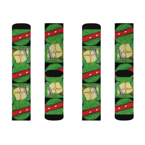 May include: Four pairs of black and green socks with a repeating pattern of a Teenage Mutant Ninja Turtle's head with a red bandana. The turtle's head is green with a red bandana and a yellow shell. The socks have the words "Pizza" and "Tacos" printed on them.