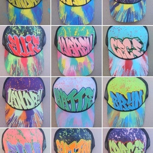May include: Twelve colorful trucker hats with different names written in graffiti style on the front. The hats are all black mesh with a variety of colors and patterns on the front.