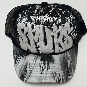 May include: A black and silver trucker hat featuring a mesh back and a foam front panel. The front displays the text SAN ANTONIO SPURS in a large, silver, graffiti-style font against a black background with white paint splatter effects.