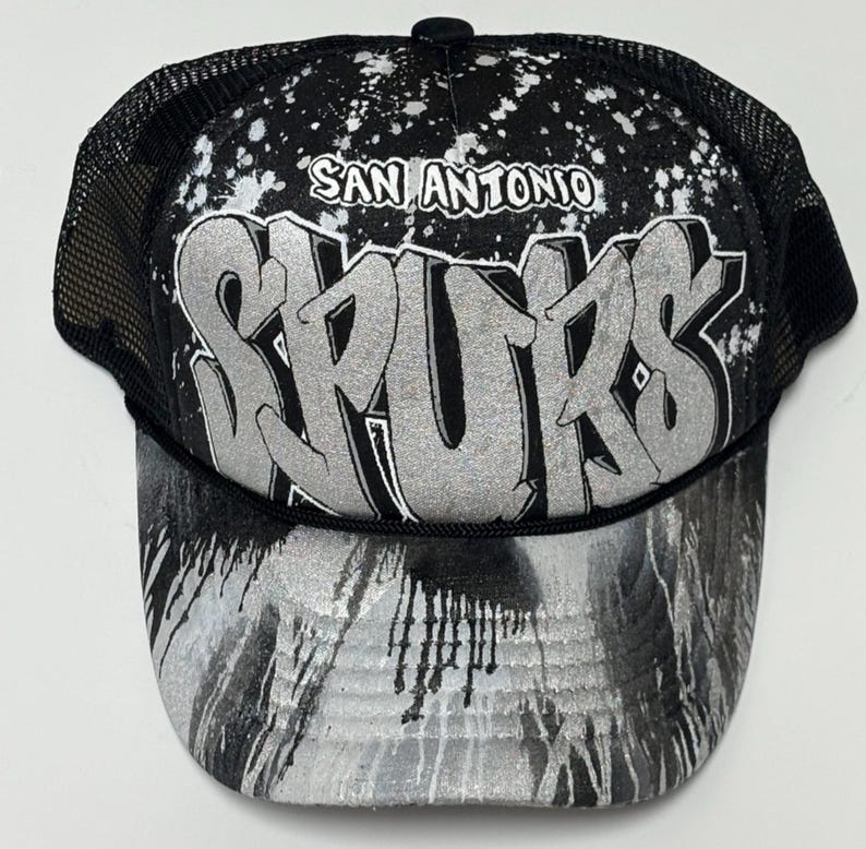 May include: A black and silver trucker hat featuring a mesh back and a foam front panel. The front displays the text SAN ANTONIO SPURS in a large, silver, graffiti-style font against a black background with white paint splatter effects.