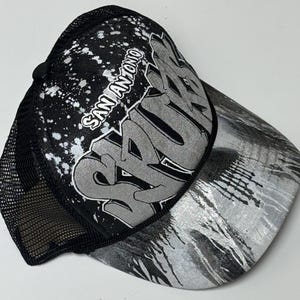 May include: A black and silver trucker hat featuring a mesh back and a foam front panel. The front displays the text San Antonio and Spurs in a stylized graffiti font, accented by a white paint splatter pattern and a silver metallic brim.