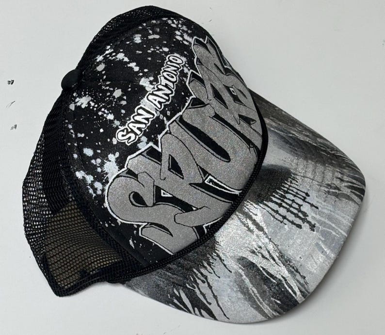 May include: A black and silver trucker hat featuring a mesh back and a foam front panel. The front displays the text San Antonio and Spurs in a stylized graffiti font, accented by a white paint splatter pattern and a silver metallic brim.