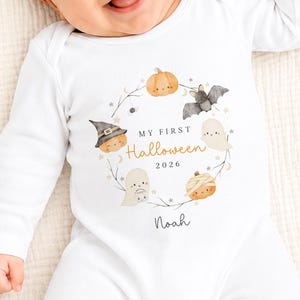 May include: A white long-sleeved baby bodysuit featuring a watercolour wreath of pumpkins, a bat, and ghosts. The central text reads My First Halloween 2026 with the name Noah printed below. The design includes small stars and moon icons in soft colours.