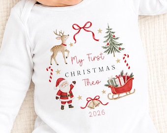 May include: A white long-sleeve baby bodysuit featuring a festive graphic with a reindeer, a Christmas tree, a red sleigh with gifts, Santa Claus, and bells. The text reads My First CHRISTMAS Theo 2026 in red and black lettering.