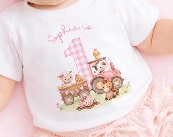 May include: A white short-sleeved t-shirt featuring a pink gingham number one, the text Sophia is, and a graphic illustration of a pink tractor with farm animals including a sheep, a cow, a pig, and a chicken in a wagon on a green field.