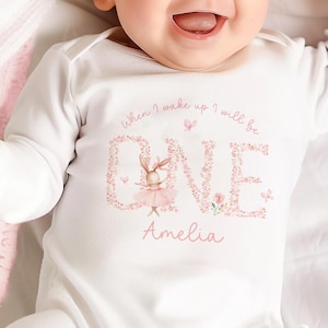 May include: A baby wearing a white long-sleeved onesie with the text "When I wake up I will be ONE" in a floral design, and the name "Amelia". The baby is smiling. A pink blanket is in the background.