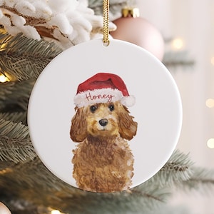 Könnte beinhalten: Runder, weißer Weihnachtsschmuck mit einer Aquarellillustration eines braunen Hundes mit roter Weihnachtsmütze. Auf der Mütze steht der Name "Honey". Der Schmuck hängt an einem goldenen Faden und befindet sich auf einem Weihnachtsbaum.