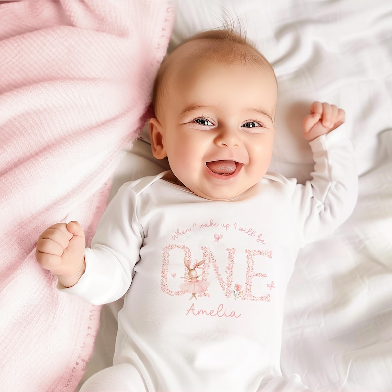 May include: A baby wearing a white long-sleeved onesie with the text "When I wake up I will be ONE" in a floral design, and the name "Amelia". The baby is smiling. A pink blanket is in the background.