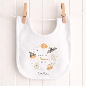 May include: A white baby bib featuring a central circular illustration of a pumpkin, a ghost, a bat, and a mummy. The text reads MY FIRST Halloween 2026 and the name Ava Mae. The bib is secured by two wooden clothes pegs to a white string.