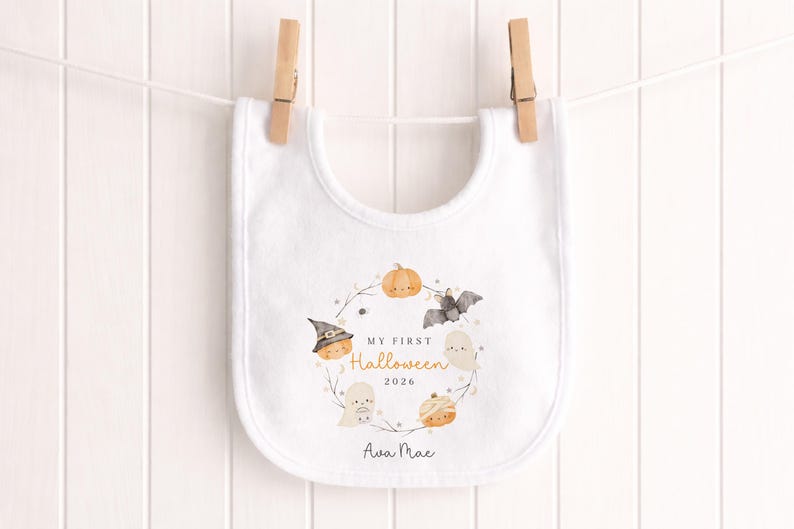 May include: A white baby bib featuring a central circular illustration of a pumpkin, a ghost, a bat, and a mummy. The text reads MY FIRST Halloween 2026 and the name Ava Mae. The bib is secured by two wooden clothes pegs to a white string.