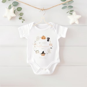 May include: White baby onesie with a Halloween-themed design. The design includes a wreath of cartoon ghosts, pumpkins, a black cat, and other spooky characters. The text "My First Halloween 2025" and a name are printed in the center.