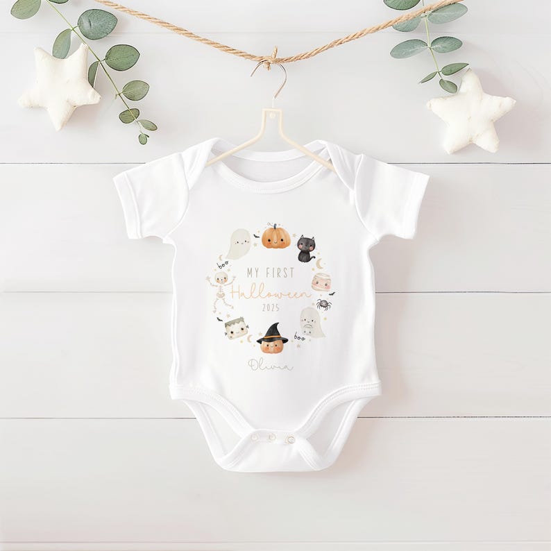 May include: White baby onesie with a Halloween-themed design. The design includes a wreath of cartoon ghosts, pumpkins, a black cat, and other spooky characters. The text "My First Halloween 2025" and a name are printed in the center.