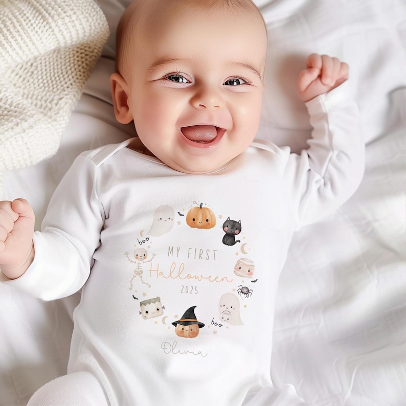 May include: A white baby onesie with a Halloween-themed design. The design includes a pumpkin, ghost, black cat, and the text "My First Halloween 2025" with the name "Olivia" below. The baby is smiling.