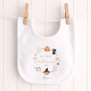 May include: A white baby bib featuring a circular illustration of Halloween characters including a pumpkin, ghost, black cat, skeleton, and spider. The text reads MY FIRST Halloween 2026 and the name Wren is printed at the bottom in a script font.