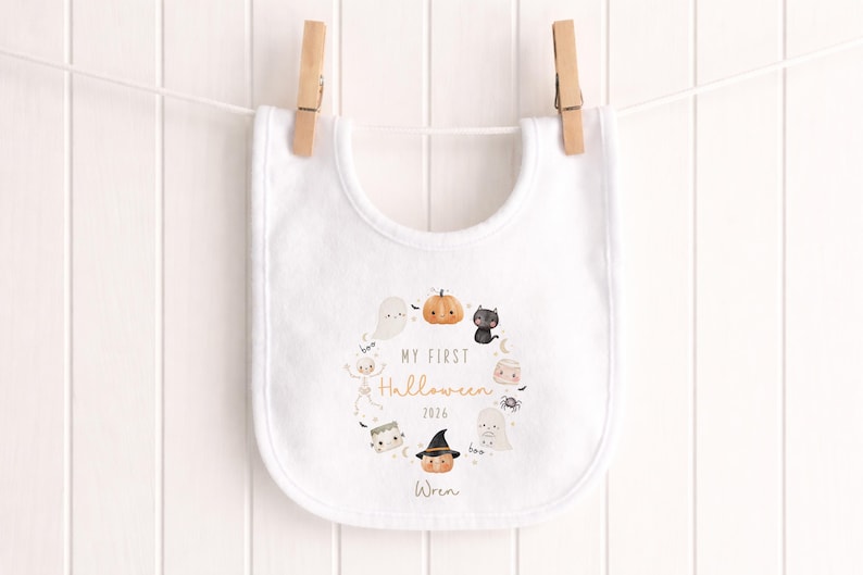 May include: A white baby bib featuring a circular illustration of Halloween characters including a pumpkin, ghost, black cat, skeleton, and spider. The text reads MY FIRST Halloween 2026 and the name Wren is printed at the bottom in a script font.