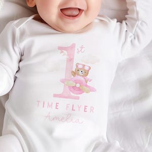 Könnte beinhalten: Weißer Baby-Langarmbody mit einer rosa Zahl 1 und einem Cartoon-Bären mit Fliegerbrille, der ein rosa Flugzeug steuert. Der Text "TIME FLYER" und ein Name sind darunter aufgedruckt. Das Baby lächelt.