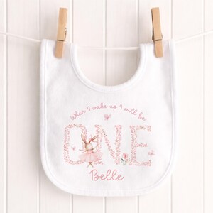 May include: White baby bib featuring a pink floral "ONE" design and a ballerina bunny illustration. The bib has the text "When I wake up I will be" above the "ONE" and the name "Belle" below. The bib is suspended on a clothesline.