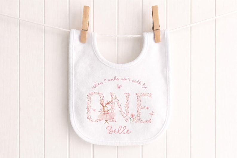 May include: White baby bib featuring a pink floral "ONE" design and a ballerina bunny illustration. The bib has the text "When I wake up I will be" above the "ONE" and the name "Belle" below. The bib is suspended on a clothesline.
