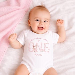 May include: A white baby bodysuit with the text "When I wake up I will be ONE Anna" in pink script and floral design. The bodysuit features a cartoon bunny wearing a tutu. The baby is lying on a white sheet with a pink blanket.