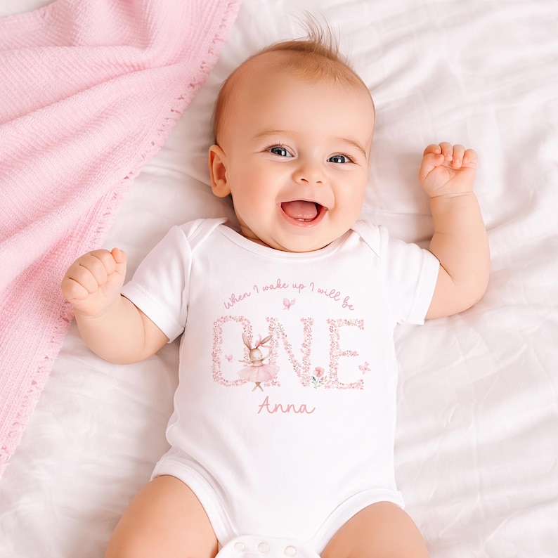 May include: A white baby bodysuit with the text "When I wake up I will be ONE Anna" in pink script and floral design. The bodysuit features a cartoon bunny wearing a tutu. The baby is lying on a white sheet with a pink blanket.