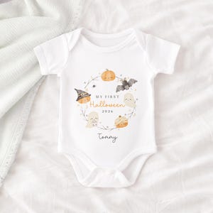 May include: A white short-sleeved baby bodysuit featuring a watercolour wreath design with pumpkins, a bat, and ghosts. The text reads "MY FIRST Halloween 2026" in the centre, with the name "Tommy" printed below the wreath in a black script font.