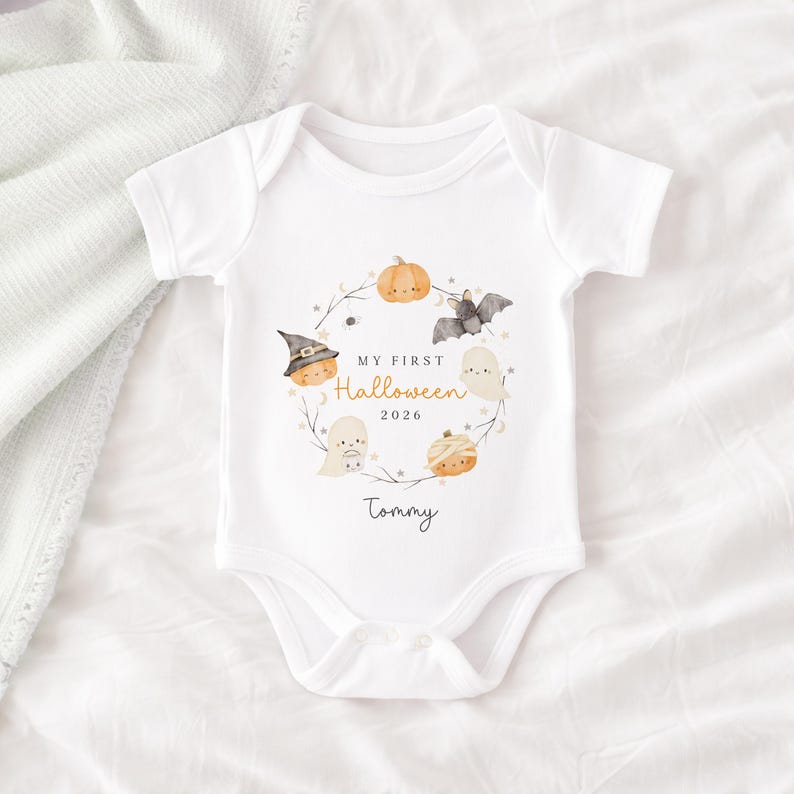 May include: A white short-sleeved baby bodysuit featuring a watercolour wreath design with pumpkins, a bat, and ghosts. The text reads "MY FIRST Halloween 2026" in the centre, with the name "Tommy" printed below the wreath in a black script font.