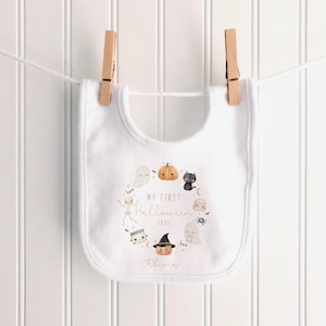 May include: White baby bib with a Halloween-themed design. The bib features a circular arrangement of cartoon illustrations including a pumpkin, ghost, and black cat. Text on the bib reads "MY FIRST HALLOWEEN 2025" and a name. The bib is hanging on a clothesline.