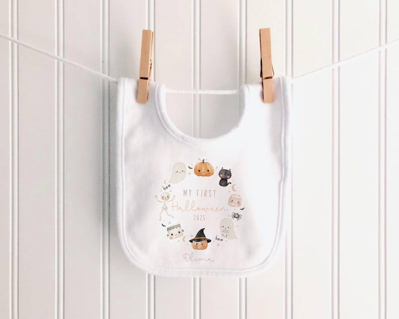May include: White baby bib with a Halloween-themed design. The bib features a circular arrangement of cartoon illustrations including a pumpkin, ghost, and black cat. Text on the bib reads "MY FIRST HALLOWEEN 2025" and a name. The bib is hanging on a clothesline.