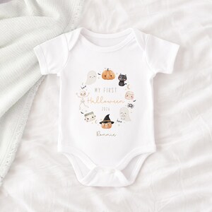 May include: A white short-sleeved baby bodysuit featuring a circular graphic of Halloween icons including a pumpkin, ghost, black cat, skeleton, and spider. The text reads My First Halloween 2026 and the name Ronnie in a script font.