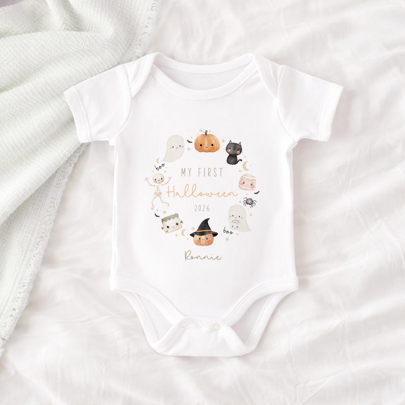 May include: A white short-sleeved baby bodysuit featuring a circular graphic of Halloween icons including a pumpkin, ghost, black cat, skeleton, and spider. The text reads My First Halloween 2026 and the name Ronnie in a script font.