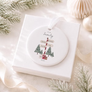 May include: A round white ceramic ornament featuring a red and white striped signpost with the text Our Family, Daddy, Mummy, Coco, and Isla, surrounded by green pine trees and wrapped gift boxes, with the year 2026 printed at the bottom.