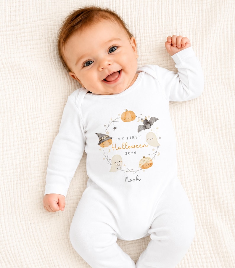 May include: A white long-sleeved baby bodysuit featuring a watercolour wreath of pumpkins, a bat, and ghosts. The central text reads My First Halloween 2026 with the name Noah printed below. The design includes small stars and moon icons in soft colours.