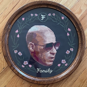 Peut inclure: Un portrait brodé au point de croix d'un homme portant des lunettes de soleil. Le portrait est entouré d'un motif floral et le mot "Famille" est écrit sous l'image.
