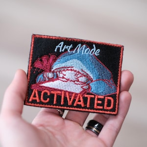 Peut inclure: Écusson brodé sur fond noir, représentant un requin stylisé en bleu, rouge et blanc. Le texte "Art Mode" est au-dessus du requin et "ACTIVATED" en dessous, tous deux en blanc et rouge. L'écusson est tenu dans une main.