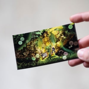 May include: A green and gold illustration of a fairy with wings and a crown, surrounded by a forest scene with glowing orbs.