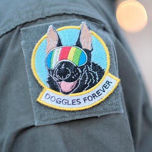 May include: A circular embroidered patch featuring a black dog wearing multicolored striped goggles against a light blue background. The patch has a yellow border and a white banner at the bottom with the black text DOGGLES FOREVER.