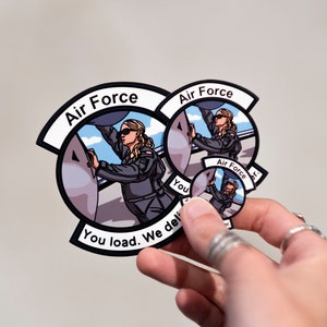 May include: Three round stickers with a cartoon illustration of a woman in an Air Force uniform loading cargo onto a plane. The largest sticker reads "Air Force" and "You load. We deliver." The medium sticker reads "Air Force" and the smallest sticker reads "Air Force".
