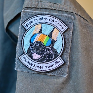 May include: A circular embroidered patch attached to an olive green fabric sleeve. The patch features a black dog wearing rainbow-colored goggles, with the text Sign in with CAC/PIV at the top and Please Enter Your Pin at the bottom.