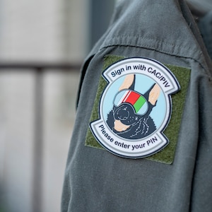 May include: A green military uniform with a patch that says "Sign in with CAC/PIV Please enter your PIN". The patch features a black dog wearing goggles with a red, white, and green flag.