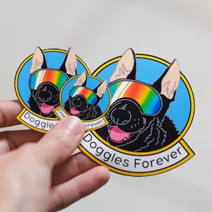 May include: Three stickers featuring a black dog wearing rainbow-colored goggles. The dog is set against a blue background with the words "Doggles Forever" in a banner. The stickers are held in a hand.