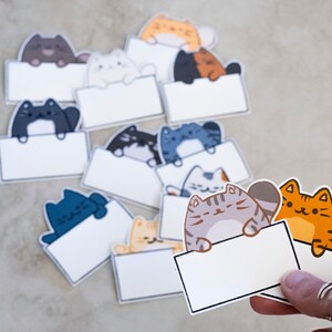 Può includere: Un set di biglietti da nota vuoti con simpatici gatti dei cartoni animati che spuntano dalla parte superiore. I gatti sono di vari colori, tra cui arancione, grigio e blu. I biglietti sono perfetti per scrivere note, promemoria o messaggi.