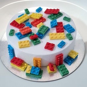 Könnte beinhalten: Ein runder weißer Kuchen, der mit zahlreichen bunten Kunststoff-Bausteinen in Rot, Blau, Gelb und Grün verziert ist. Die Bausteine sind auf der Oberseite verstreut und vertikal an der Seite des Kuchens angeordnet.
