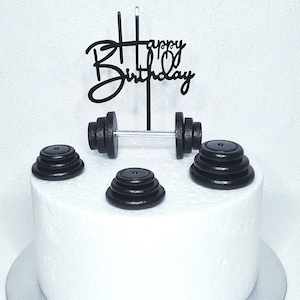Könnte beinhalten: Ein weißer Kuchen, verziert mit schwarzen Hantelscheiben und einem "Happy Birthday"-Tortentopper. Der Kuchen hat eine runde Form und steht auf einem silbernen Kuchenbrett. Die Hantelscheiben sind um den Kuchen herum angeordnet.
