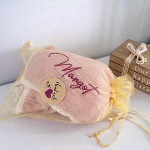 May include: A soft pink fabric item with a cream-colored lace trim, enclosed in a sheer gold organza drawstring bag. The fabric features the embroidered name Margot in a dark pink script and a round sticker reading Handmade with a purple heart.