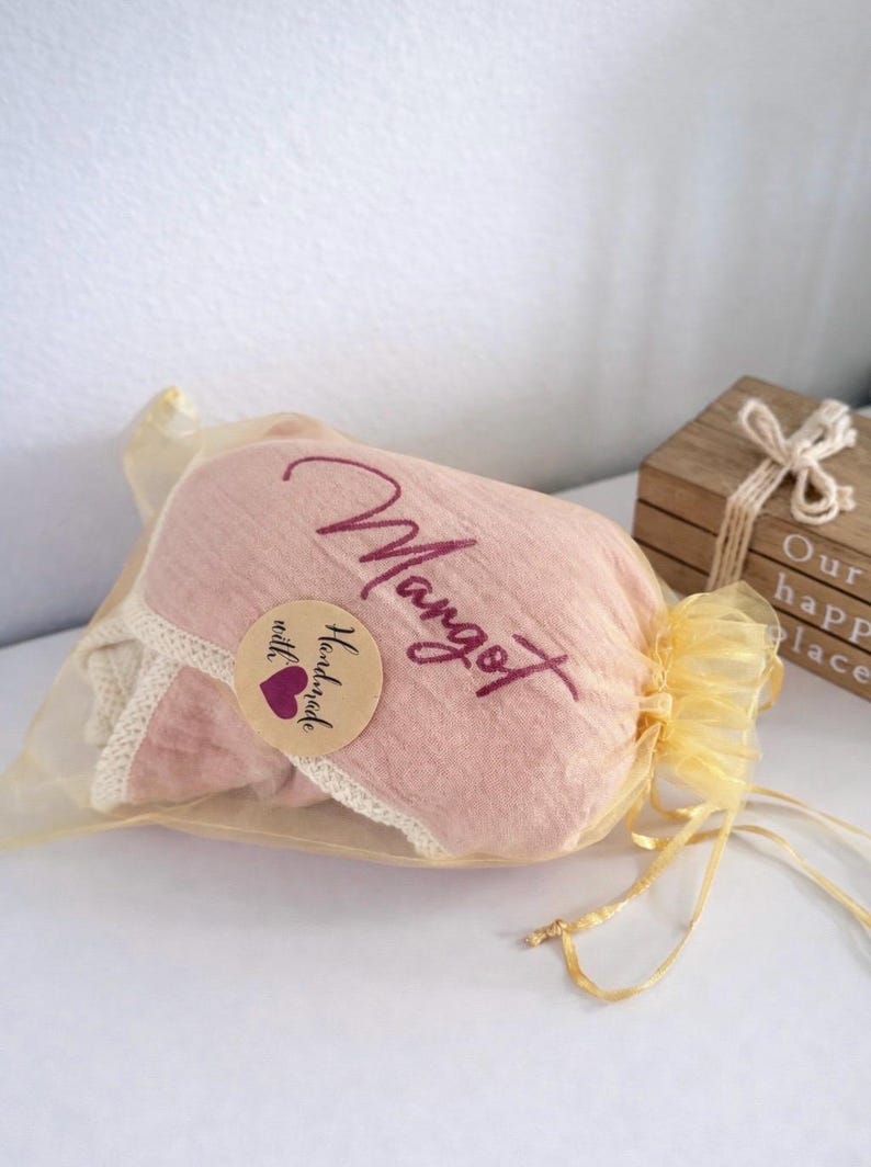 May include: A soft pink fabric item with a cream-colored lace trim, enclosed in a sheer gold organza drawstring bag. The fabric features the embroidered name Margot in a dark pink script and a round sticker reading Handmade with a purple heart.