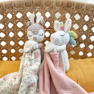 May include: Two crochet security blankets featuring a deer and a unicorn. The deer has a beige head with antlers and a floral print blanket. The unicorn has a white head with a rainbow horn and mane, paired with a solid light pink muslin blanket.